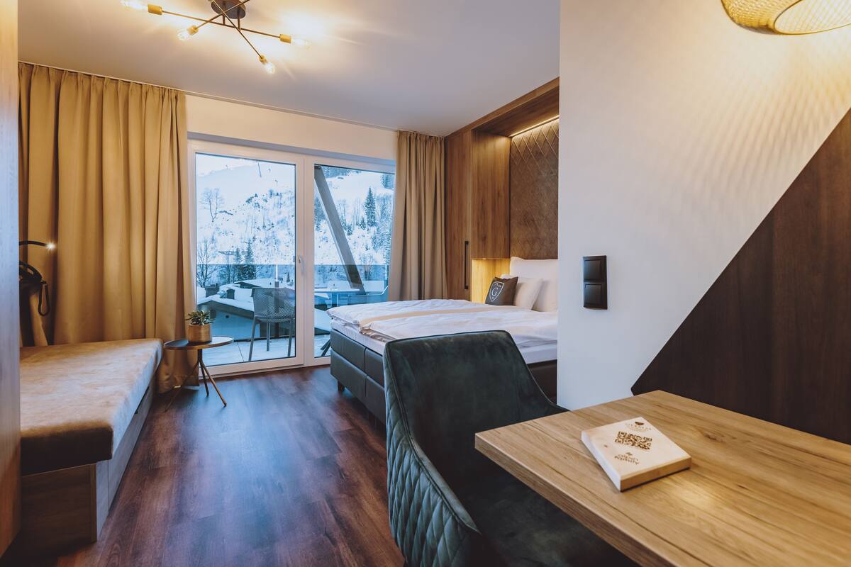 Ein modernes Hotelzimmer mit einem Bett, einem Schreibtisch, einem Sessel, einem kleinen Tisch und Blick auf einen verschneiten Berg.