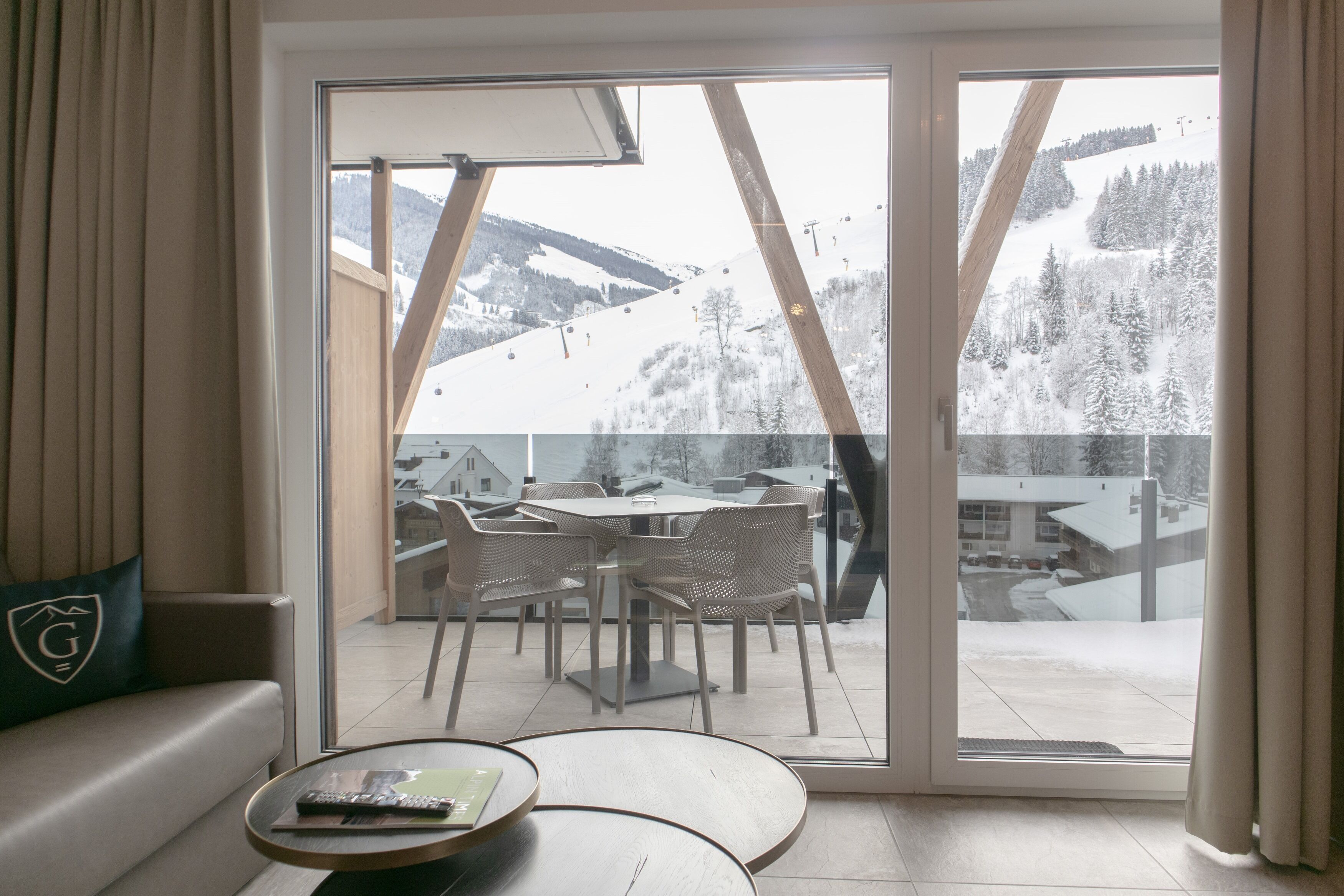 A room with a large sliding glass door leading to a balcony with a view of a snowy mountain and ski lift. Inside, there is a sofa with a cushion featuring a logo, a small round table with a remote control, and a chair.