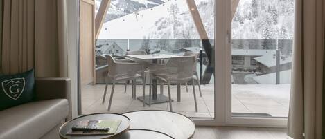 A room with a large sliding glass door leading to a balcony with a view of a snowy mountain and ski lift. Inside, there is a sofa with a cushion featuring a logo, a small round table with a remote control, and a chair.