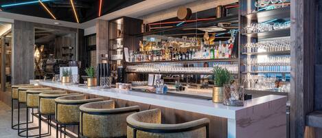 A modern bar area with a long counter, multiple stools, and shelves stocked with bottles and glasses.