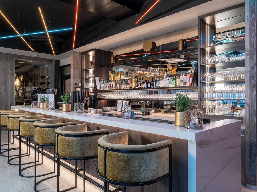 Een moderne bar met een lange toonbank, meerdere barkrukken en flessen en glazen op de planken.