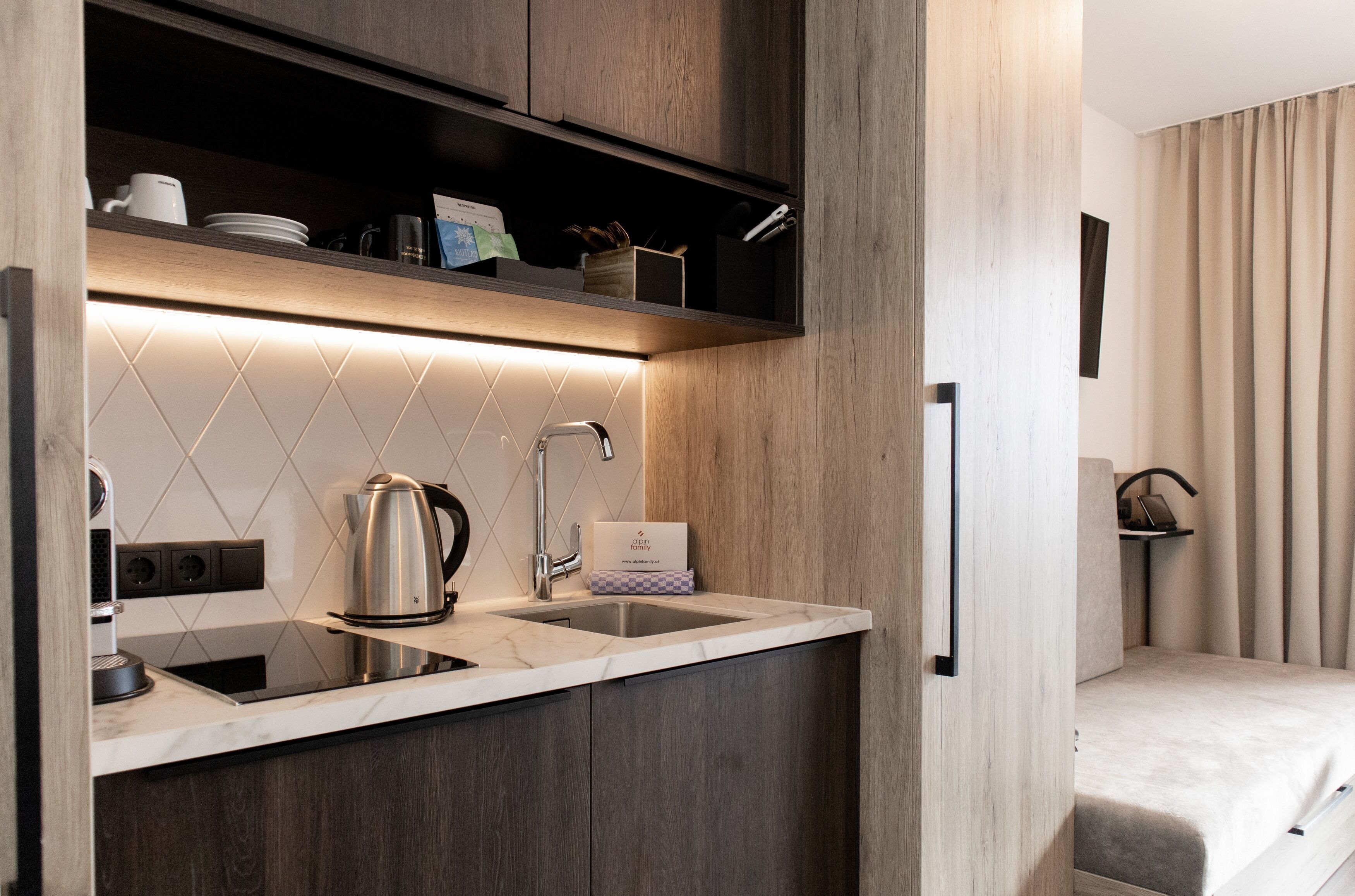 A compact kitchen with a built-in sink, a kettle, and a coffee maker. There is a shelf above the sink with dishes and a box.