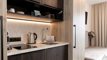 A compact kitchen with a built-in sink, a kettle, and a coffee maker. There is a shelf above the sink with dishes and a box.