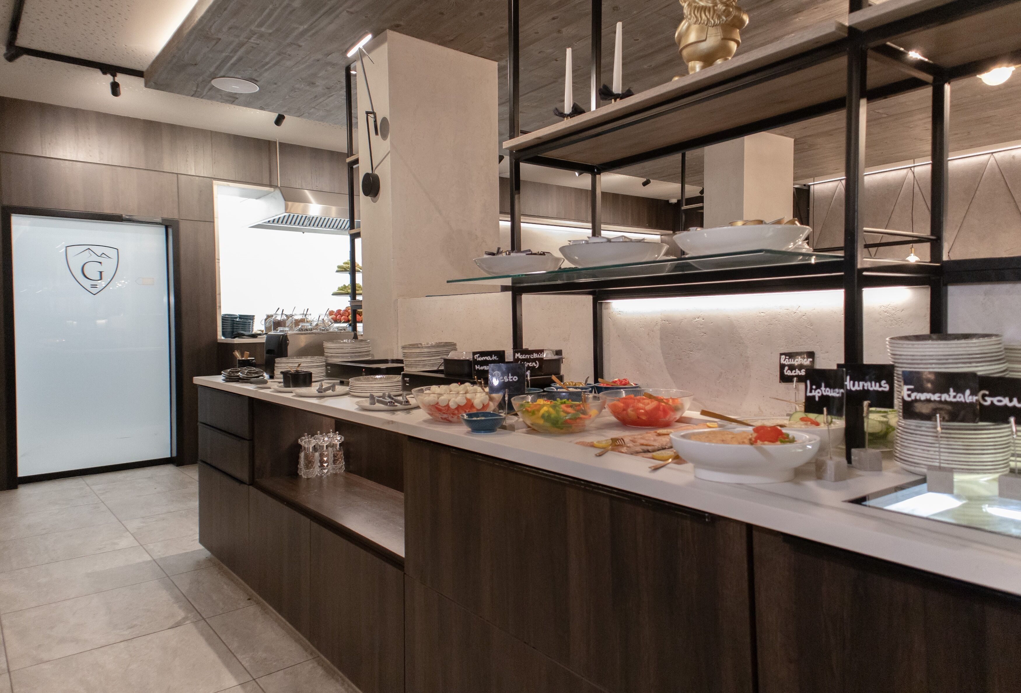 A modern buffet setup with various food stations labeled, a glass display case, and a logo on the door.