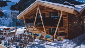 A wooden chalet with a sloped roof, outdoor seating area, and a view of a snowy mountain. - GLEMM by AvenidA Superior Hotel & Residences (Saalbach-Hinterglemm)