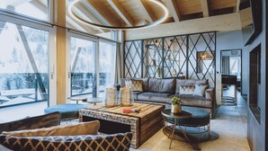 A modern living room with a wooden ceiling, large windows, a leather sofa, and a coffee table. - GLEMM by AvenidA Superior Hotel & Residences (Saalbach-Hinterglemm)