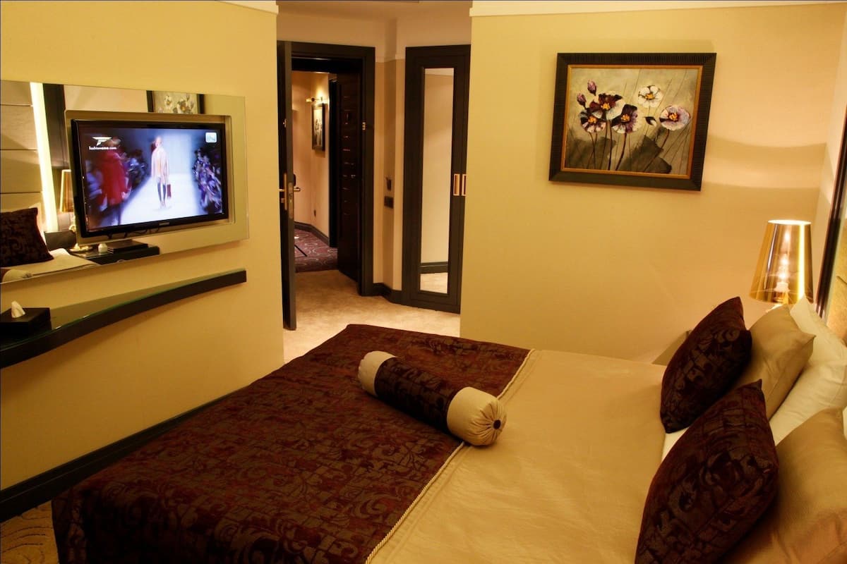 Quarto de hotel com cama, televisão exibindo um desfile de moda, quadro na parede e corredor que dá para outro quarto.