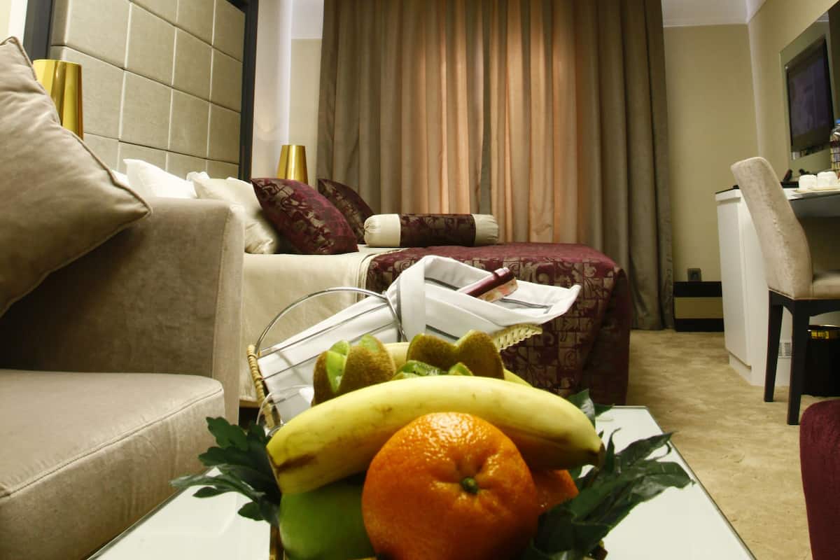 Quarto de hotel com design moderno, incluindo um sofá, uma mesa de jantar com frutas, uma escrivaninha e uma janela grande com cortinas.