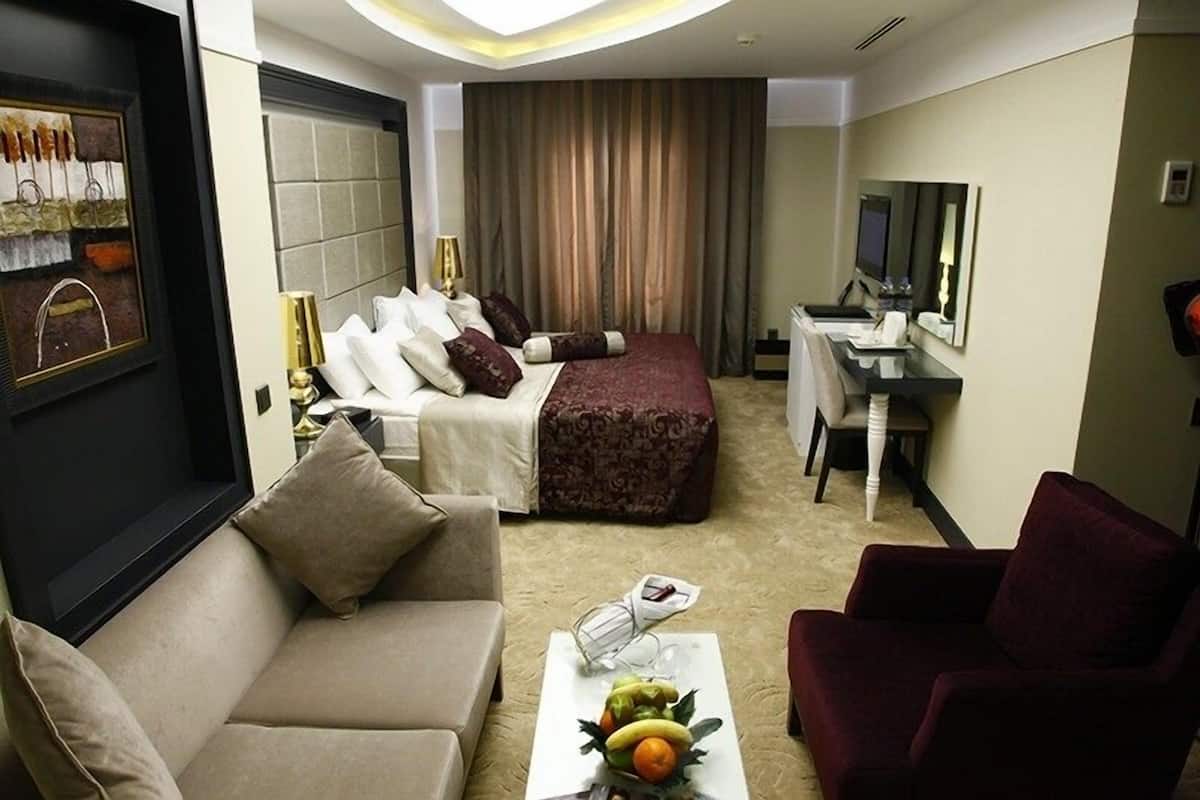Quarto de hotel com uma cama, duas mesas de cabeceira, uma escrivaninha, uma cadeira, um sofá e uma mesa de centro com frutas.