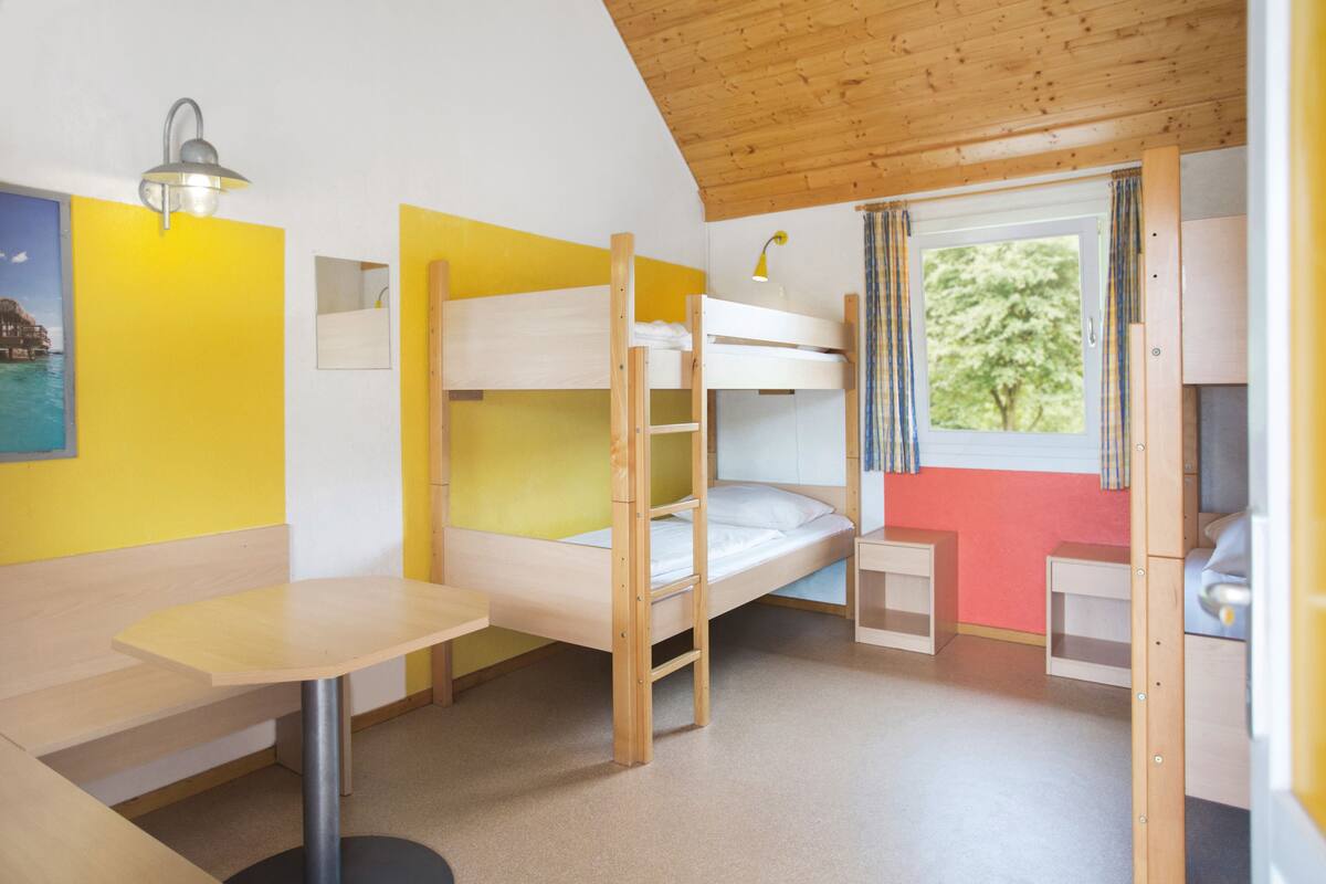 A bunk bed room with a wooden ceiling, a small table, and a window with curtains.