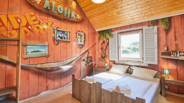 A room with a wooden bed, a hammock, and a wall with 'ALOHA' and 'HAVE A GREAT DAY' decorations. - Heide Park Holiday Camp (Soltau)