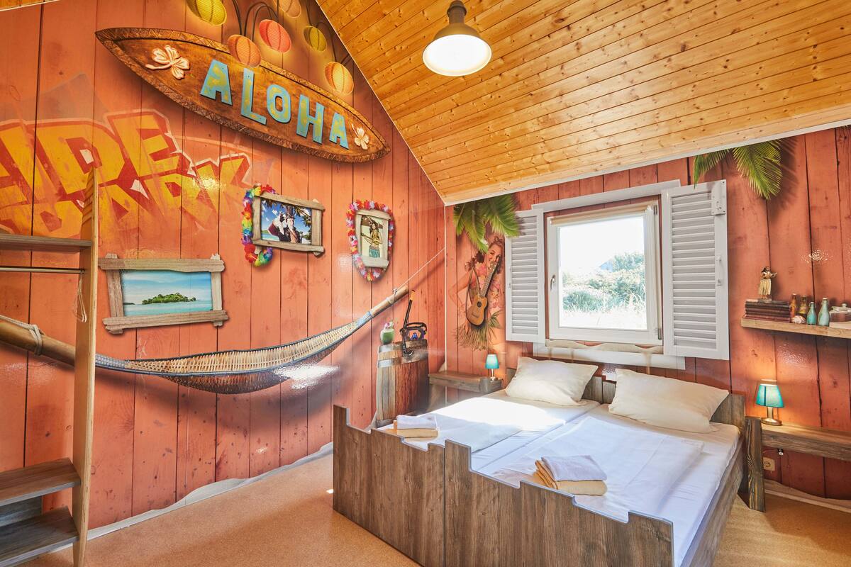 A room with a wooden bed, a hammock, and a wall with 'ALOHA' and 'HAVE A GREAT DAY' decorations.