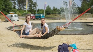 Two people sitting in a hammock on a sandy beach with a water fountain in the background. - Heide Park Holiday Camp (Soltau)