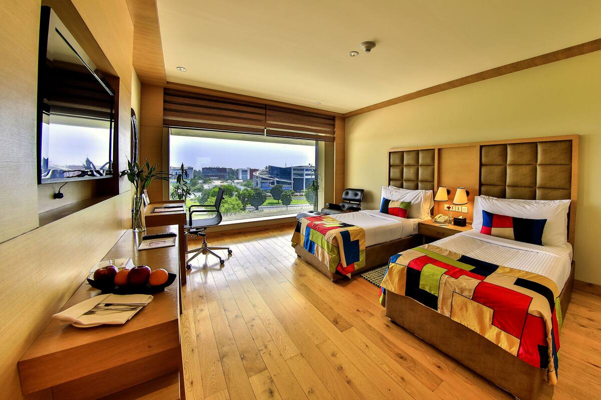 A hotel room with two beds, a large window with a city view, a desk with a chair, and a dining table with fruits and a napkin.