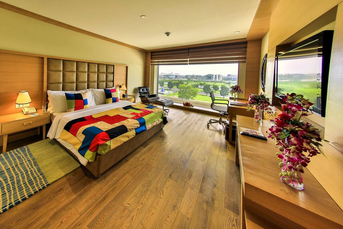 A modern hotel room with a large bed, a desk, and a view of a green landscape.