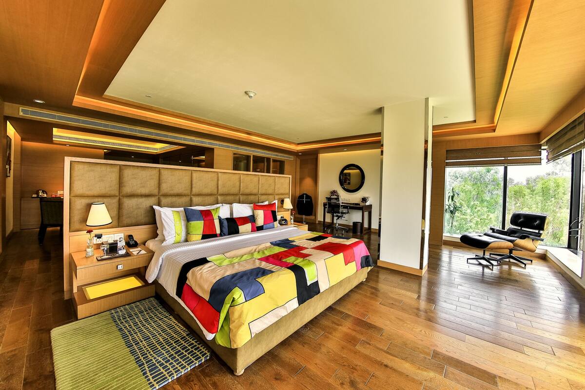 A modern hotel room with a large bed, a desk, and a view of greenery through large windows.
