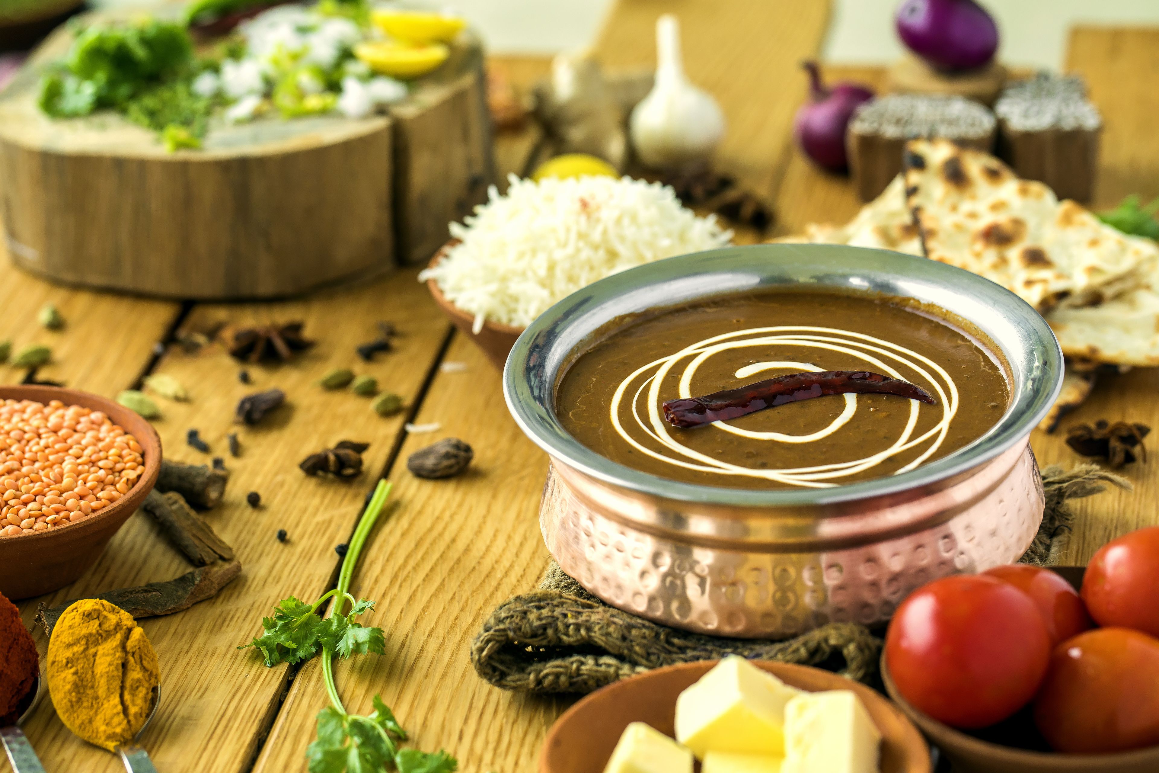 A traditional Indian meal with a metal bowl of dal, surrounded by various ingredients like tomatoes, onions, and spices.