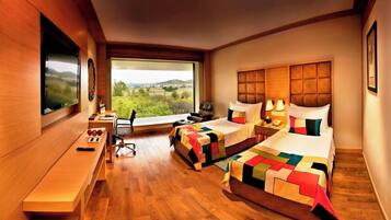 A hotel room with two beds, a desk, a TV, and a large window with a view of greenery.