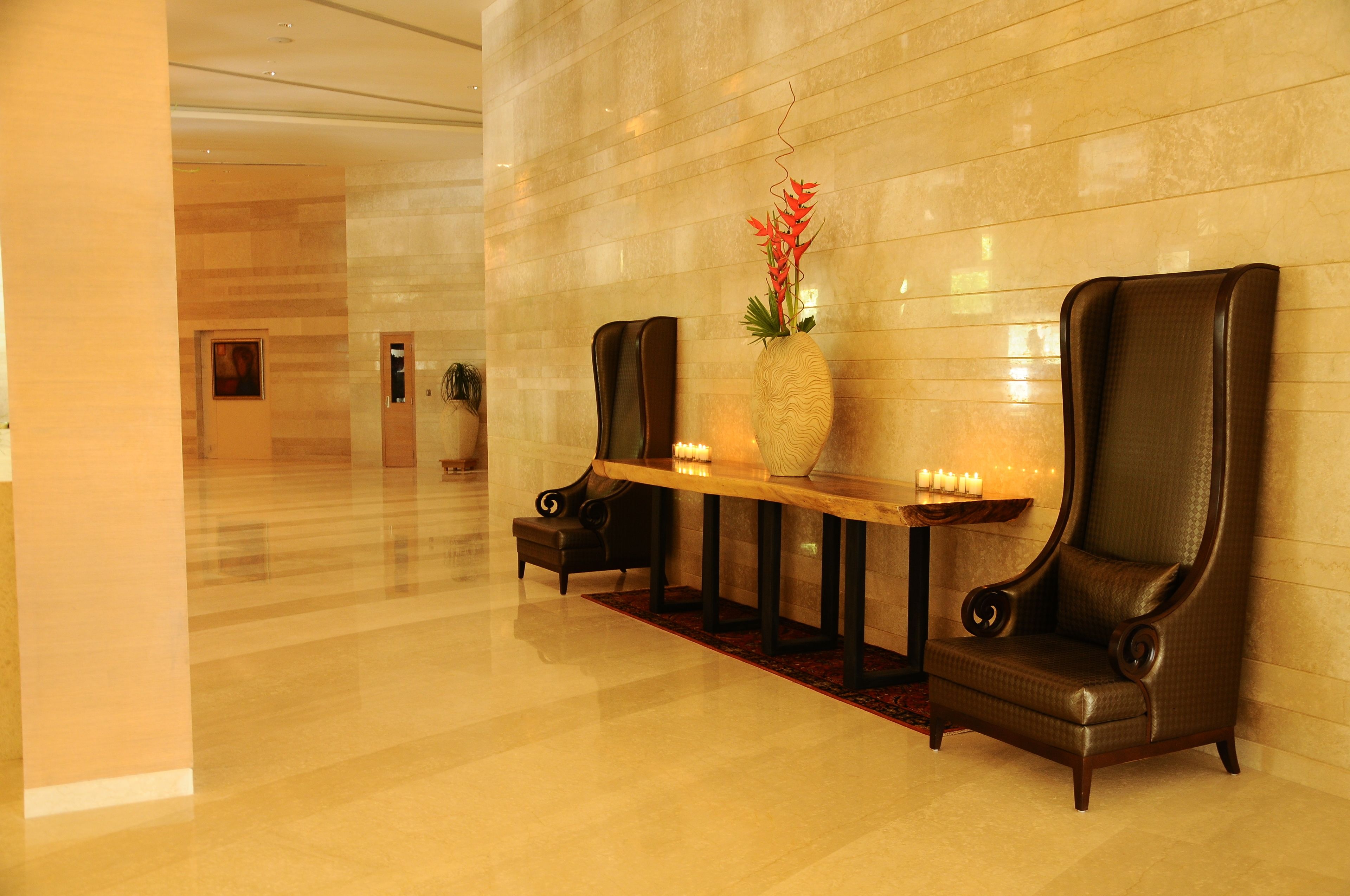 A lobby with a polished floor, a reception desk, two leather armchairs, a vase with red flowers, and a wall-mounted light fixture.