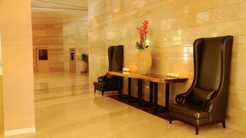 A lobby with a polished floor, a reception desk, two leather armchairs, a vase with red flowers, and a wall-mounted light fixture.