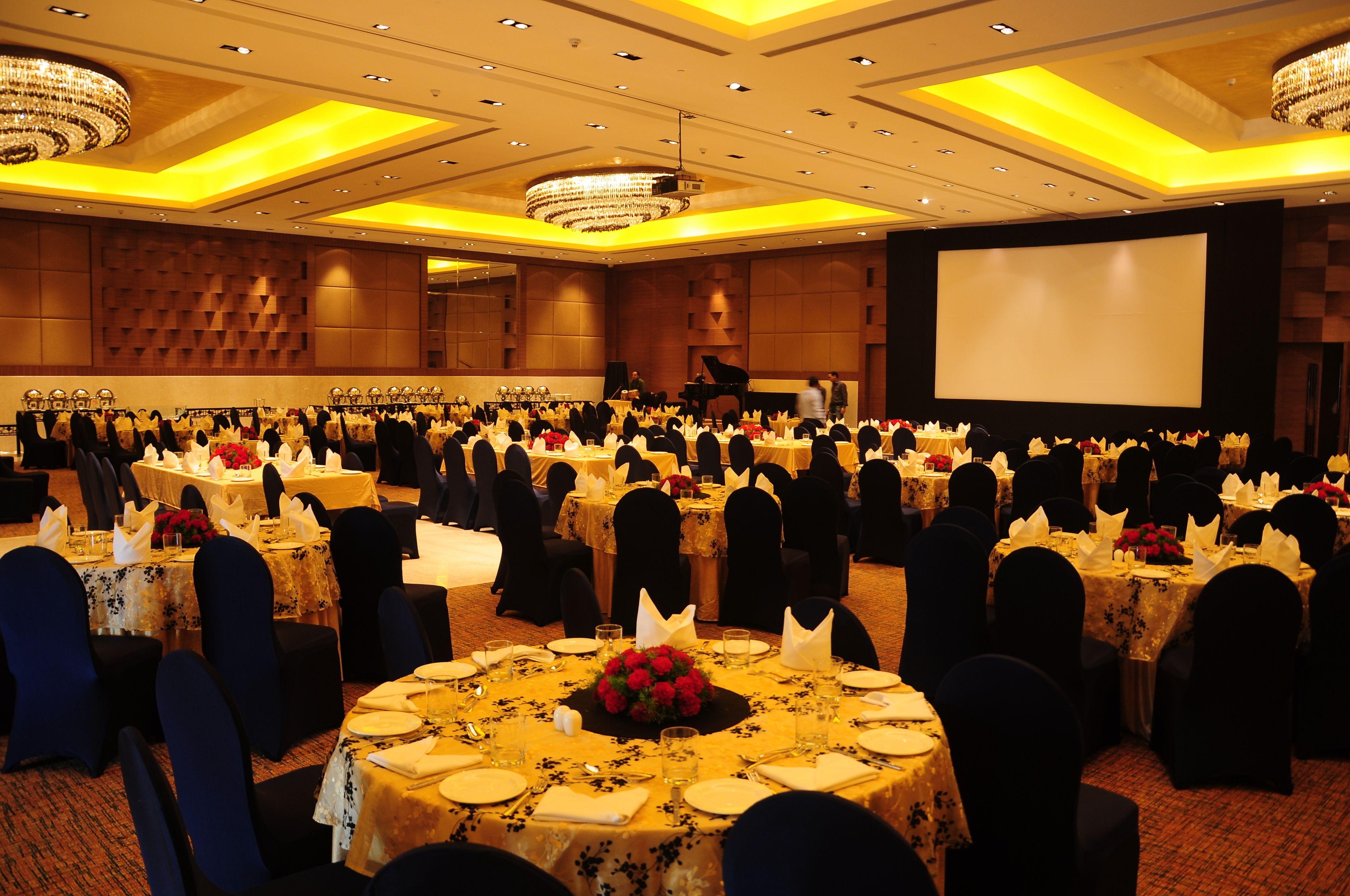 Un amplio salón de banquetes con mesas redondas dispuestas para un evento formal, un escenario con un piano y una gran pantalla.