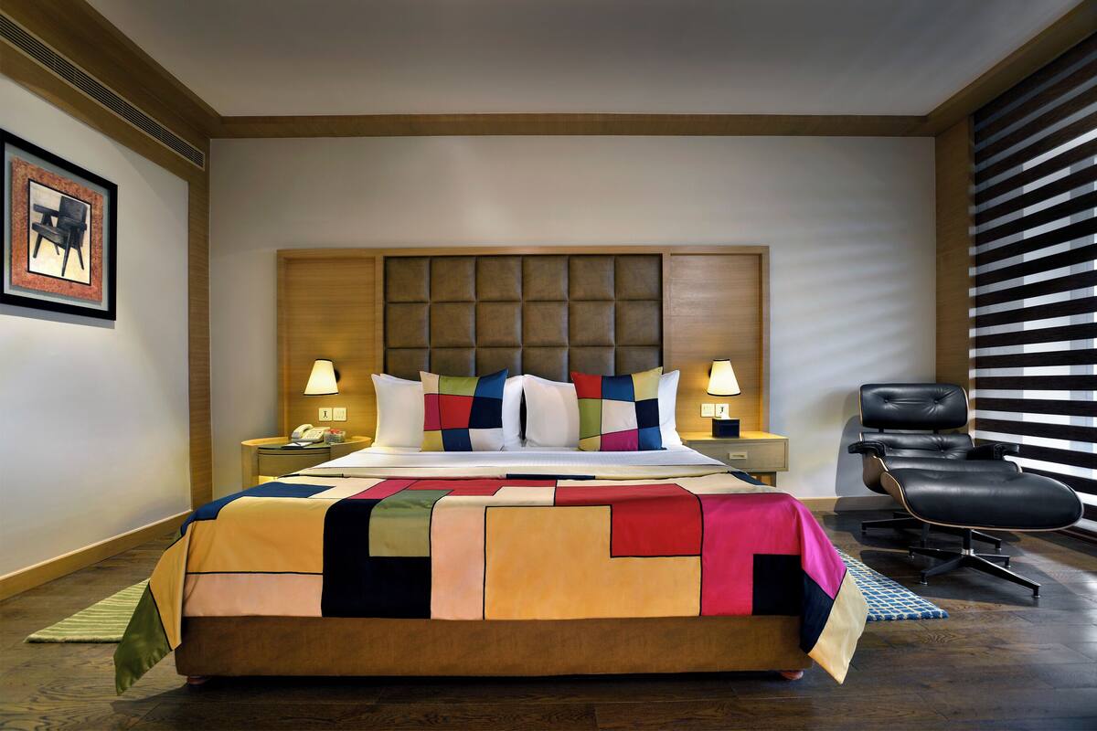 A hotel room with a large bed, a headboard, two bedside tables, a chair, and a window with blinds.