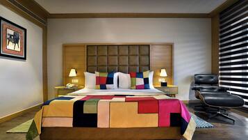 Habitación de hotel con una cama grande, cabecera, dos mesitas de noche, una silla y una ventana con persianas.
