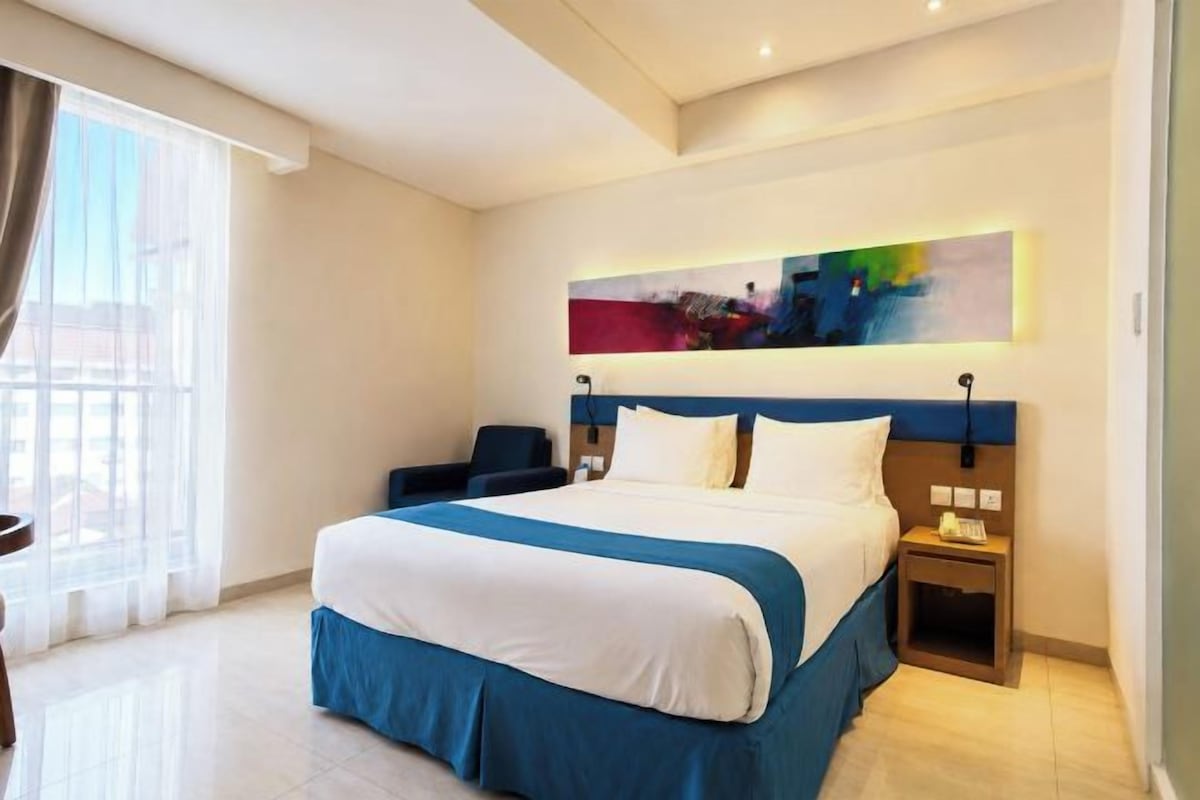 A hotel room with a large bed, a desk, a chair, a nightstand, and a painting on the wall.