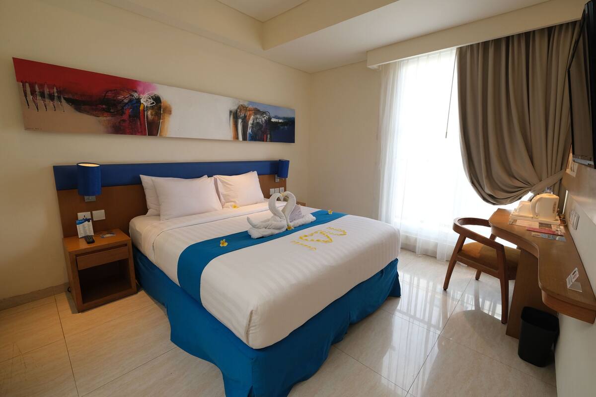 A hotel room with a bed, a desk, a chair, a nightstand, a lamp, and artwork on the wall.