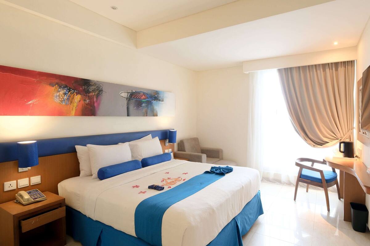 A hotel room with a bed, bedside tables, a desk, a chair, a TV, and a painting on the wall.