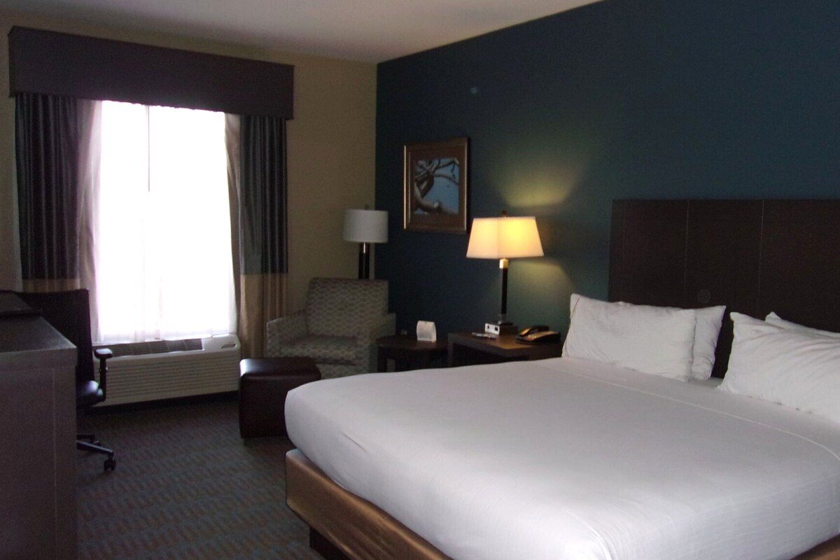 A hotel room with a large bed, a chair, a desk with a lamp, and a window with curtains.