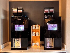 A coffee station with two espresso machines, a display of Costa Coffee branded products, and a wooden shelf with disposable cups. - Holiday Inn Express Hotel & Suites Goldsboro - Base Area by IHG (Goldsboro)