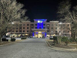 Edifício do Holiday Inn Express & Suites com entrada central, vários espaços de estacionamento e árvores ao redor.