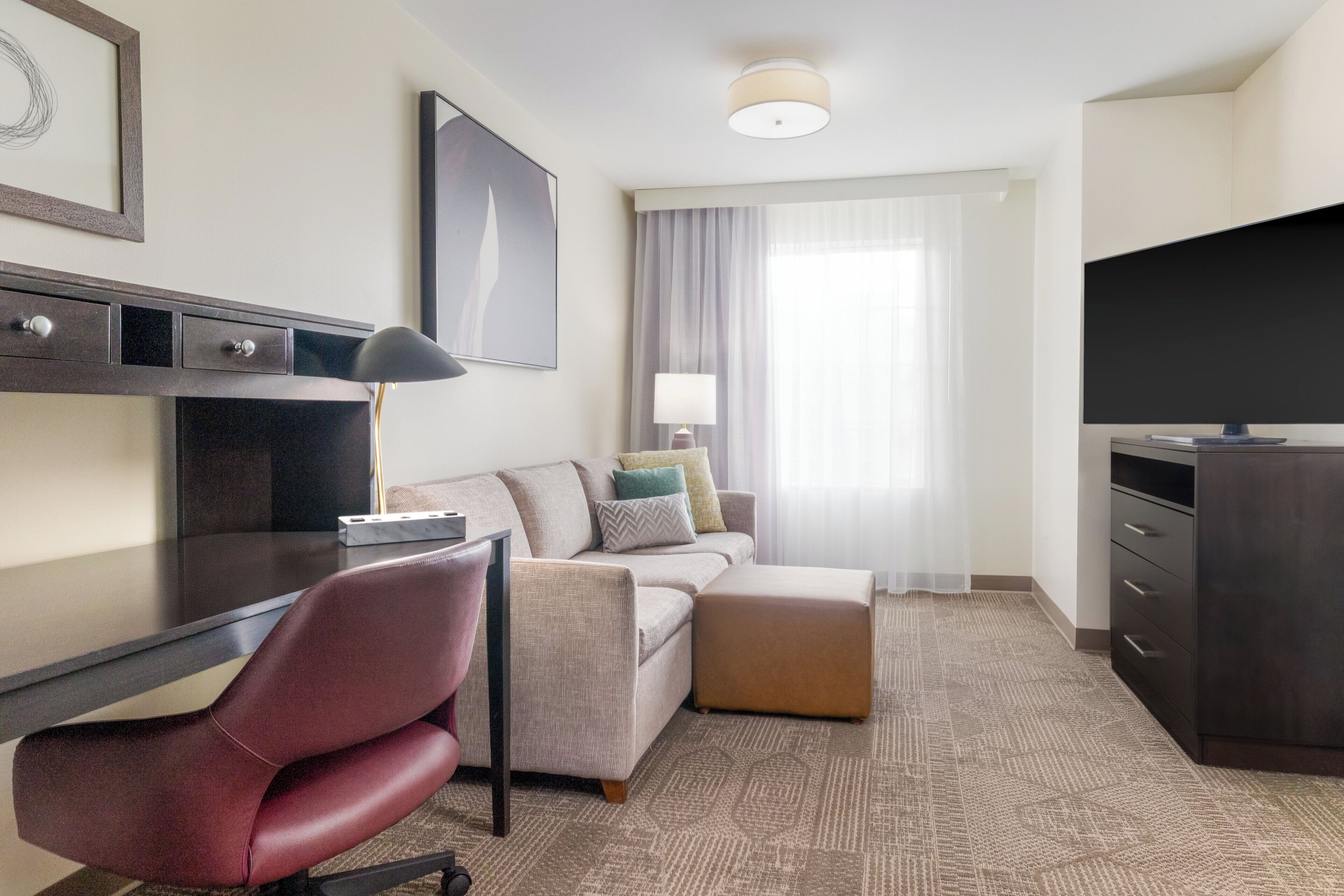 Une chambre d’hôtel équipée d’un bureau, d’un fauteuil rouge, d’un canapé, d’un pouf, d’une télévision et d’une grande fenêtre avec des rideaux.