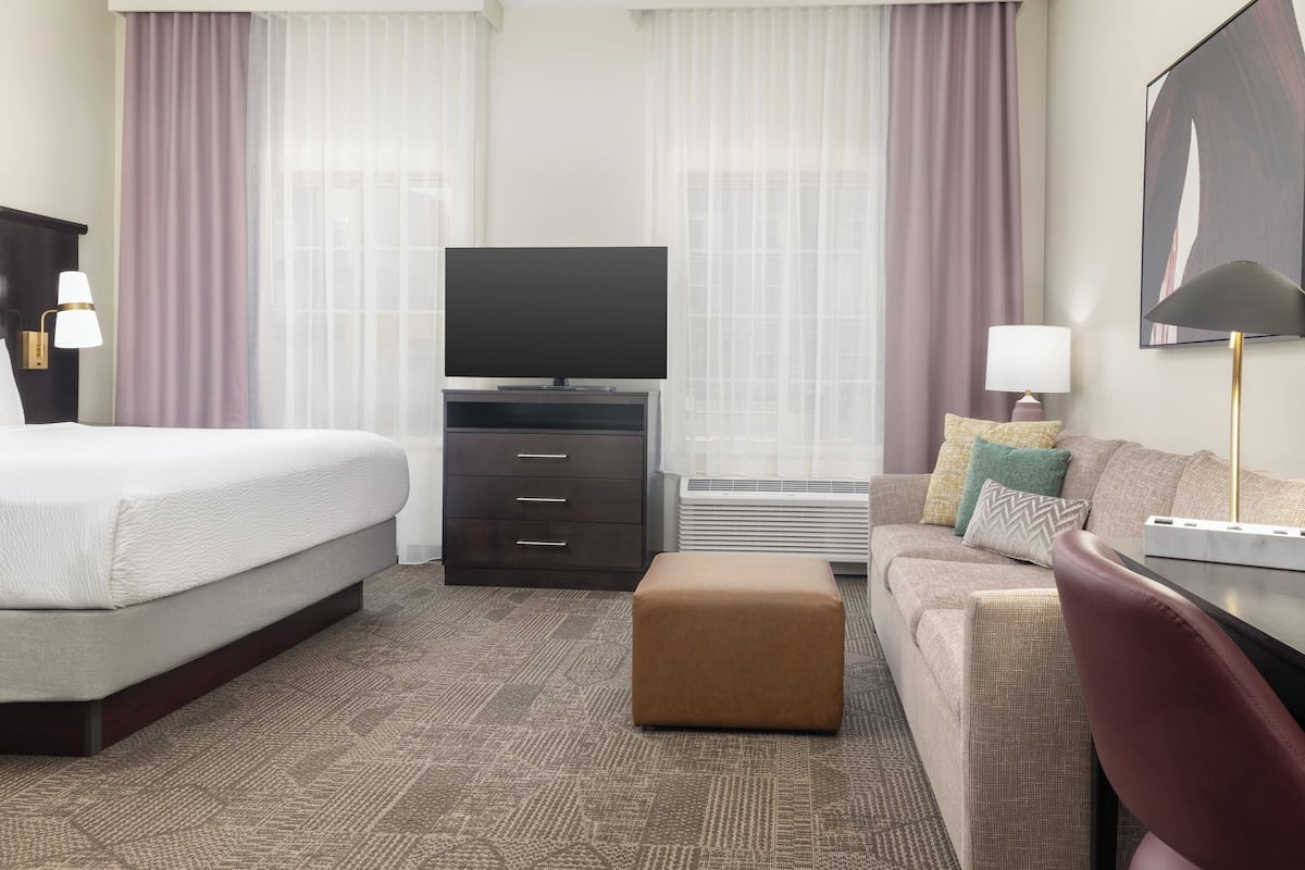 Una habitación de hotel con cama, televisor, sofá, escritorio y ventana con cortinas.