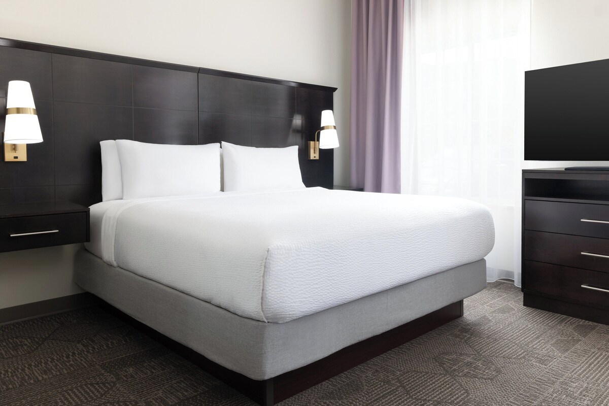 A hotel room with a large bed, dark headboard, two bedside lamps, a flat-screen TV, and a dark wooden dresser.