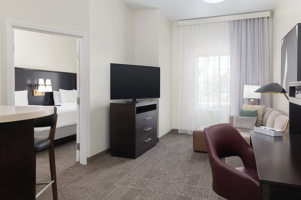 Una habitación de hotel con cama, escritorio, televisión, silla y ventana con cortinas.