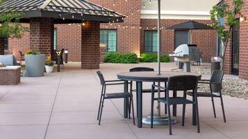 An outdoor patio area with a round table and chairs, a pergola with string lights, and a brick building in the background.
