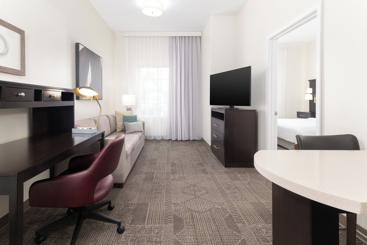 A hotel room with a dark wood desk, a red office chair, a sofa, a TV on a stand, a dining table, and a large window with sheer curtains.