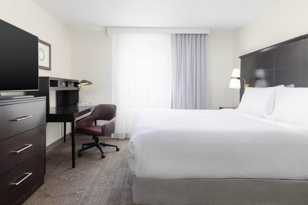 A hotel room with a bed, a desk with a chair, a TV, and a lamp.