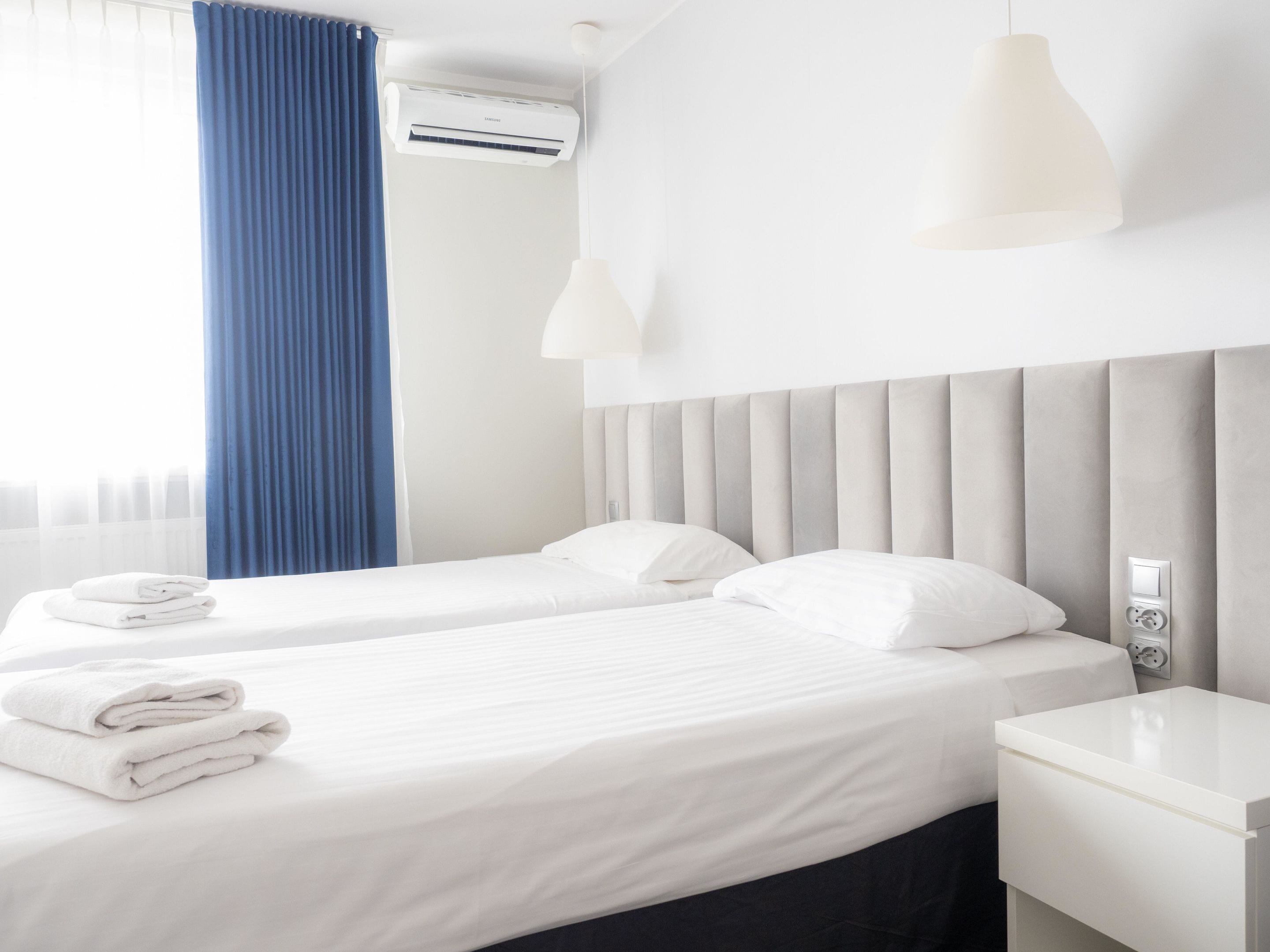 Ein Hotelzimmer mit zwei Betten, einem weißen Nachttisch, einer Wand-Klimaanlage und einem Fenster mit blauen Vorhängen.