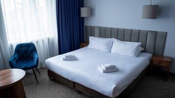 A hotel room with a large bed, a blue armchair, a round wooden table, and a window with curtains.