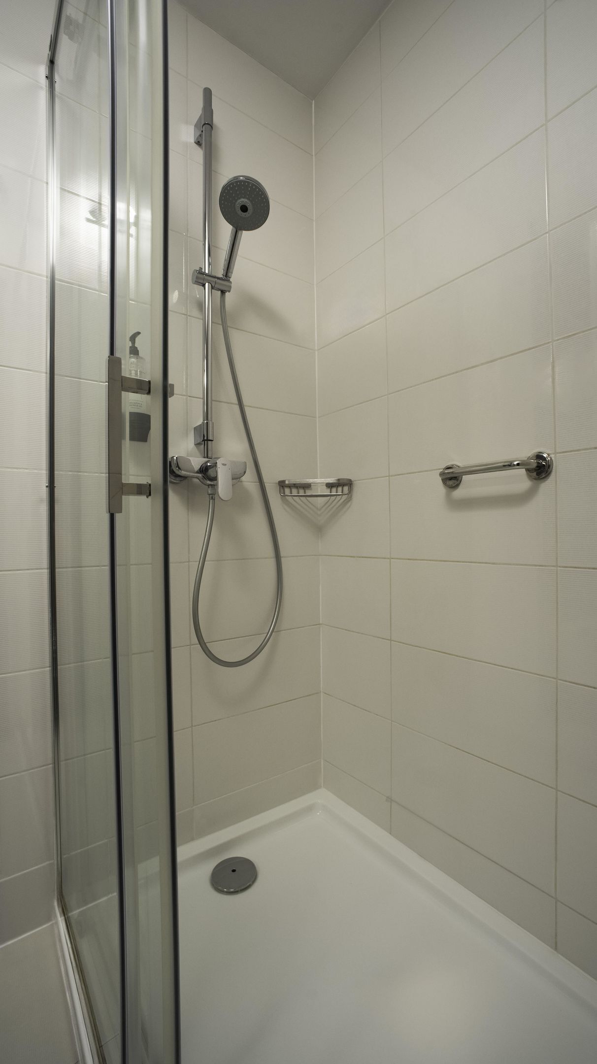 A shower with a glass door, a handheld showerhead, a handheld shower attachment, a soap dispenser, and a small shelf.