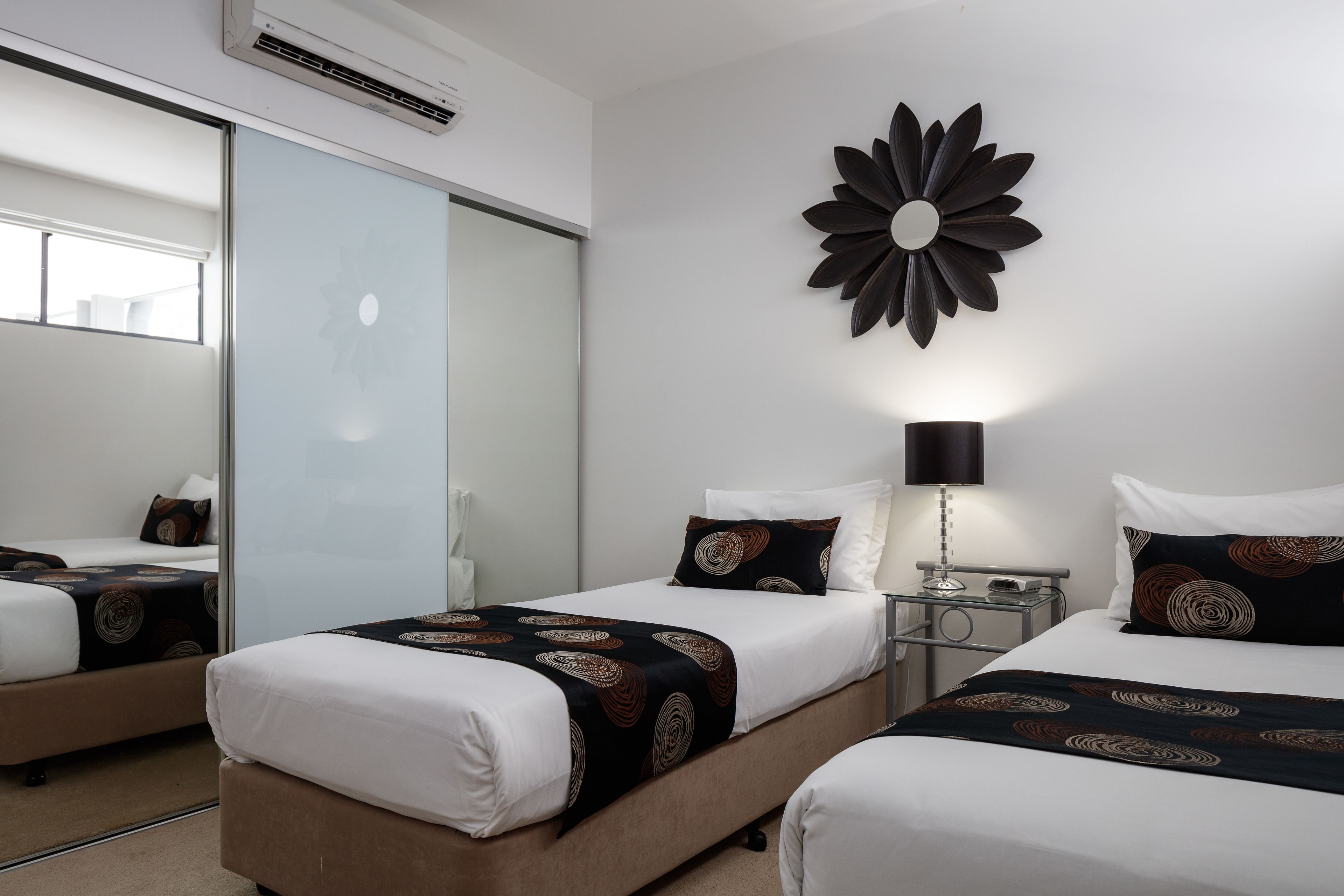 Habitación de hotel con dos camas, un espejo grande, un adorno floral en la pared y una mesita de noche con lámpara.
