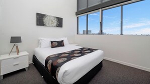 A bedroom with a large window, a bed with a patterned bedspread, a nightstand with a lamp, and a wall-mounted artwork. - Islington at Central (Townsville)