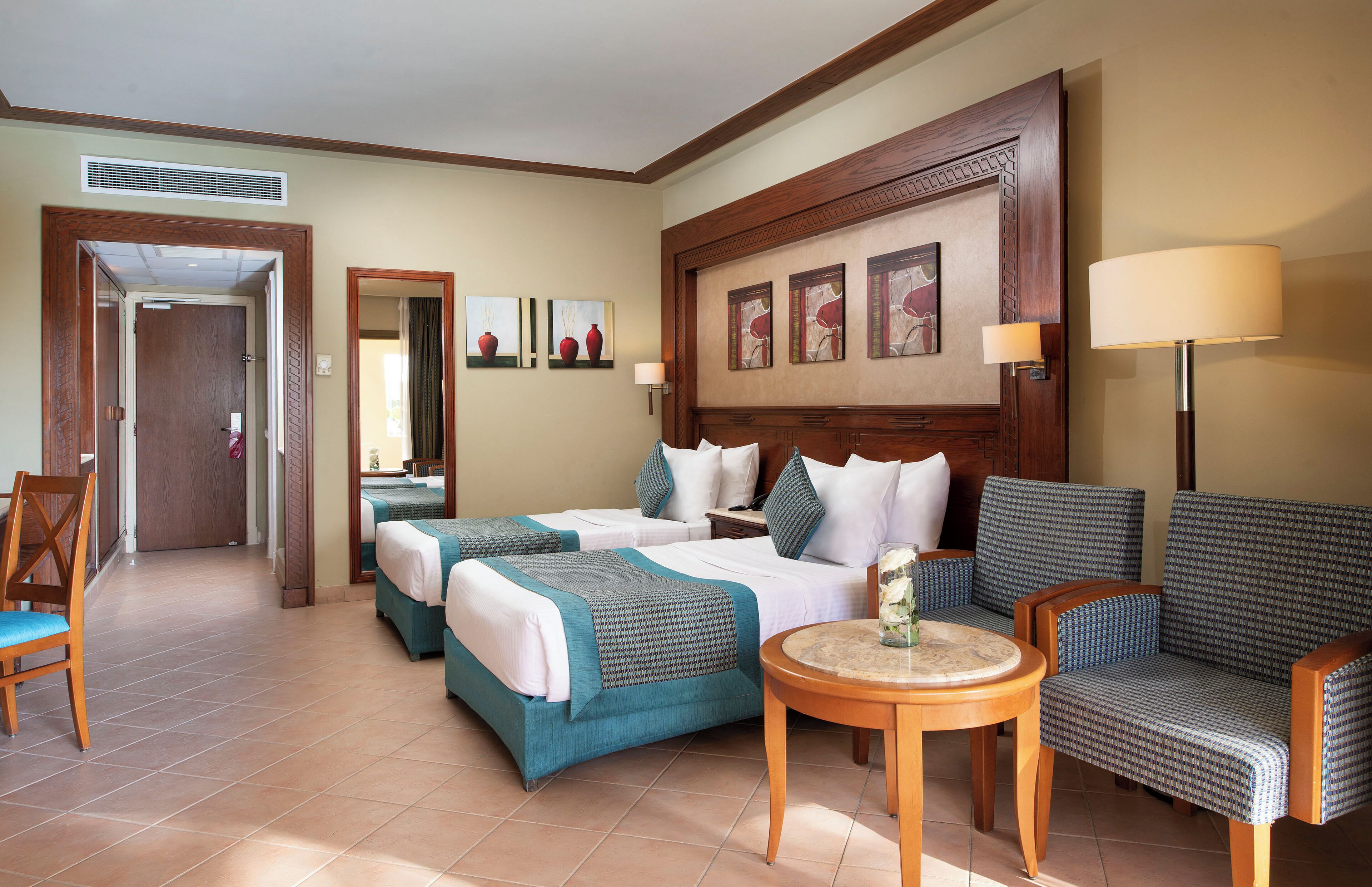 A hotel room with two beds, a sofa, a small table, and a chair. There is a door leading to another room and a window with curtains.