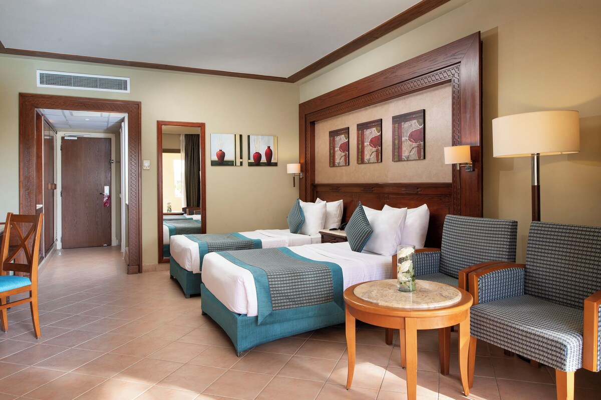 A hotel room with two beds, a sofa, a small table, and a chair. There is a door leading to another room and a window with curtains.