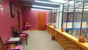 A room with a pink wall, wooden furniture, a window with a view of a building, and a wooden door. - Kurumi (Cusco)