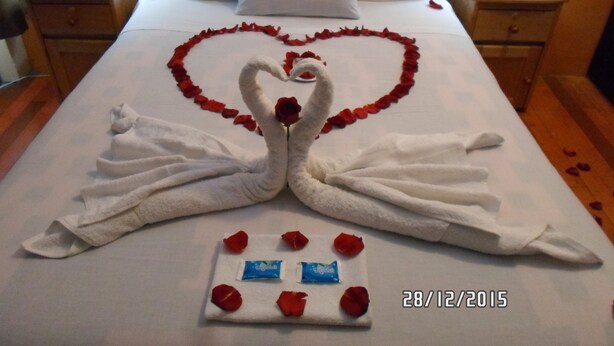 A hotel room with a bed featuring a heart-shaped arrangement of rose petals and two swan-shaped towels. There are two bedside tables with lamps, and a small tray with rose petals and chocolates on the bed. - Kurumi (Cusco)