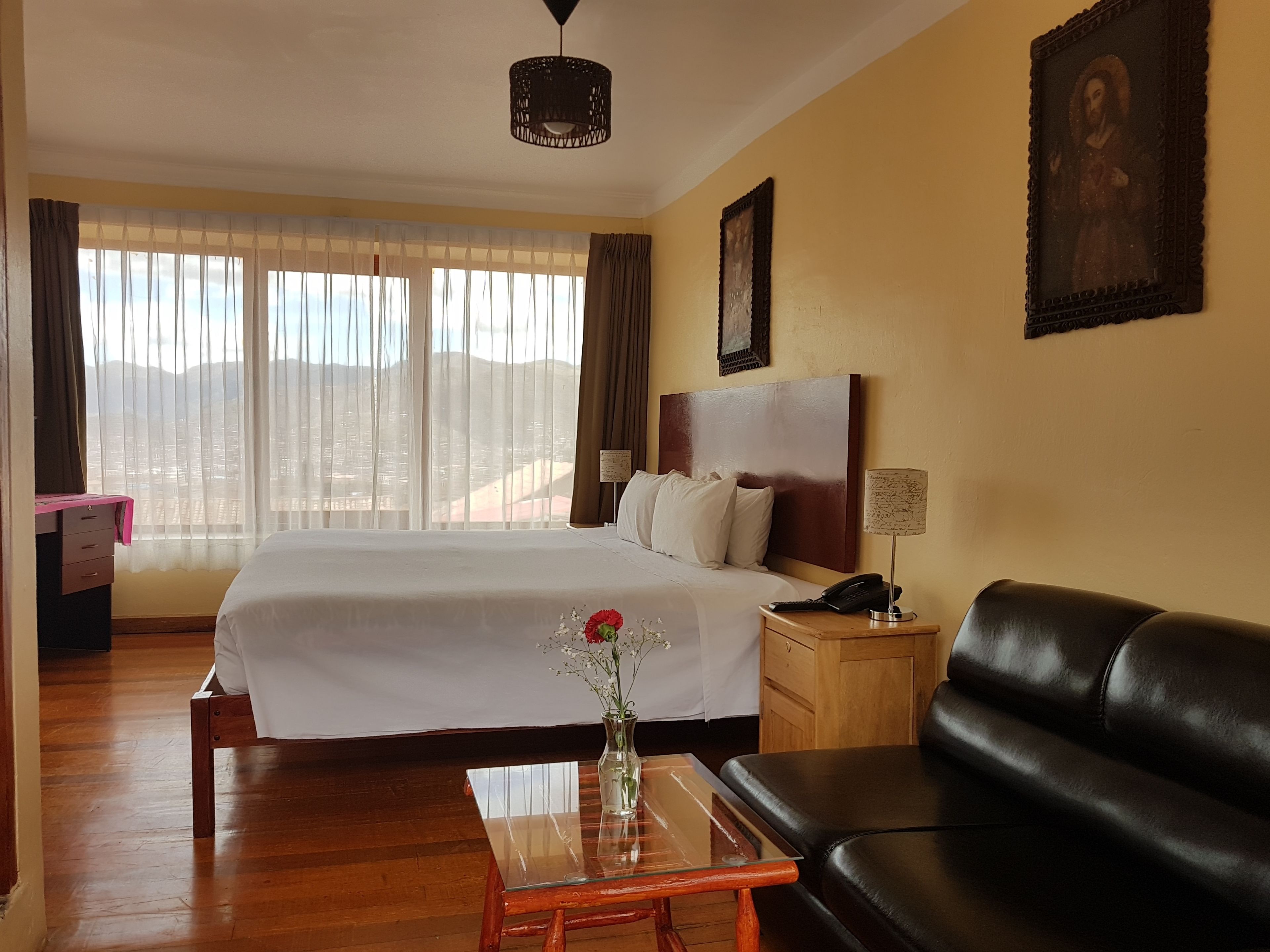 A hotel room with a large bed, a wooden nightstand, a black leather sofa, a glass coffee table, and a view of the mountains through the window.